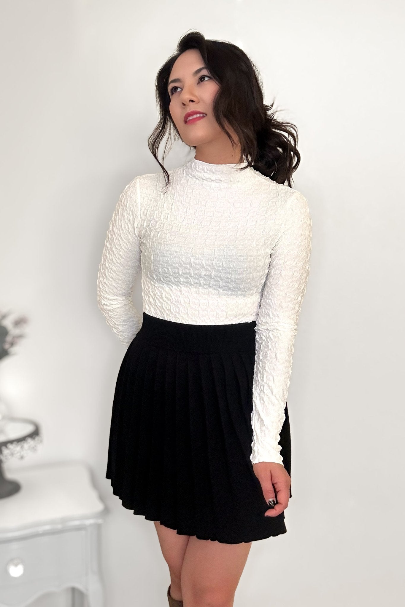 Woman wearing an off-white textured mock neck shirt and a black pleated mini skirt