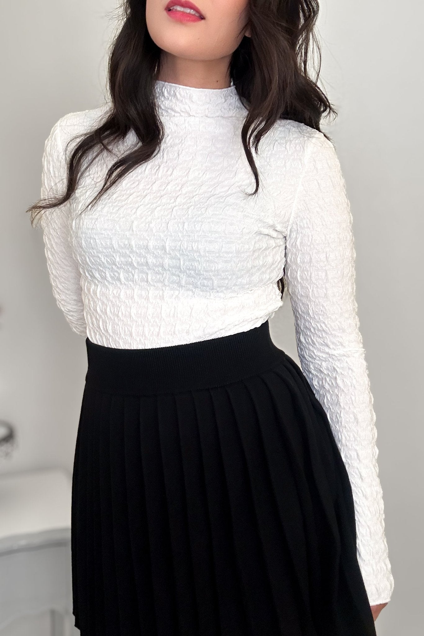 Torso view of a woman wearing an off-white textured mock neck shirt and a black pleated mini skirt