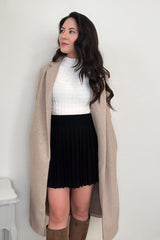 Woman wearing an off-white textured mock neck shirt and a black pleated mini skirt and an oat-colored coat